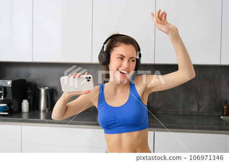 Portrait of beautiful sports woman, wearing activewear, dancing in kitchen with headphones, listening music and having fun, feeling energy after workout session 106971346