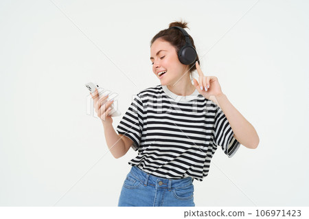 Portrait of happy woman with smartphone changes song on mobile phone streaming app, listens music in headphones, dancing against white background 106971423