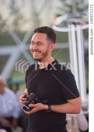 Close-up portrait of a young stylish man with a beard and dark hair in a black T-shirt. Male photographer uses a mirrorless camera to take a picture. Close-up portrait of a young stylish man with a beard and dark hair in a black T-shirt. Male photographer uses a mirrorless camera to take a picture. 106972272