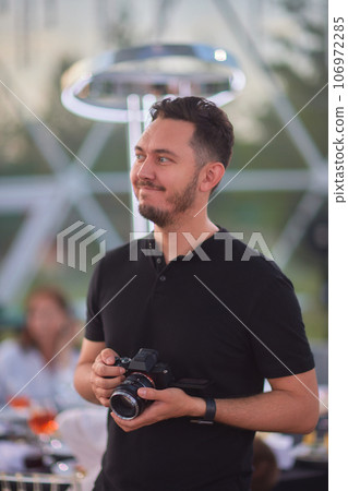 Close-up portrait of a young stylish man with a beard and dark hair in a black T-shirt. Male photographer uses a mirrorless camera to take a picture. Close-up portrait of a young stylish man with a beard and dark hair in a black T-shirt. Male photographer uses a mirrorless camera to take a picture. 106972285