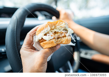 Asian lady holding hamburger to eat in car, dangerous and risk an accident. 106973093