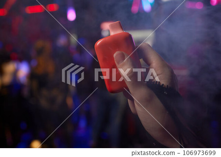 Caucasian woman smokes disposable vape on black background. Alternative device for smoking. Close-up of a female hand. Caucasian woman smokes disposable vape on black background. Alternative device for smoking. Close-up of a female hand. 106973699