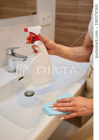 Cropped image of beautiful young woman using a duster and a detergent while cleaning sink and tap in bathroom. Cropped image of beautiful young woman using a duster and a detergent while cleaning sink and tap in bathroom. 106974569