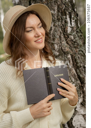 A young beautiful woman in a dress and hat stands and reads a Bible book in nature. Romantic and vintage photo of a beautiful girl. The book has the Bible written on it. Faith in God concept 106975687