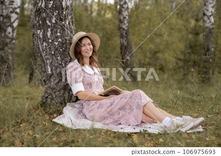 A young beautiful woman in a dress and a round hat reads a book outdoors in the forest and drinks tea. Romantic and vintage photo of a beautiful girl. Reading and relaxation 106975693