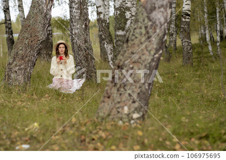 A young beautiful woman in a dress and a round hat reads a book outdoors in the forest and drinks tea. Romantic and vintage photo of a beautiful girl. Reading and relaxation A young beautiful woman in a dress and a round hat reads a book outdoors in the forest and drinks tea. Romantic and vintage photo of a beautiful girl. Reading and relaxation 106975695