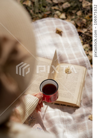 A young beautiful woman in a dress and a round hat reads a book outdoors in the forest and drinks tea. Romantic and vintage photo of a beautiful girl. Reading and relaxation A young beautiful woman in a dress and a round hat reads a book outdoors in the forest and drinks tea. Romantic and vintage photo of a beautiful girl. Reading and relaxation 106975832