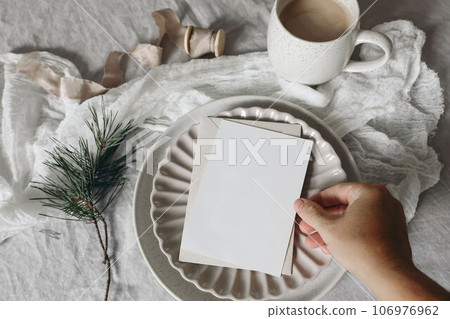 Christmas still life. Female hand holding blank vertical greeting card, invitation mockup. Scalloped plate. Pne tree branches, silk ribbon. White linen tablecloth. Cup of coffee, winter breakfast. Top 106976962