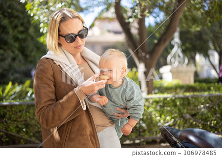 Close up portrait of sweet toddler kid eating fruit puree from plastic doy pack, sitting in stroller, outdoor snack time 106978485