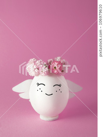Smiling angel in a wreath of flowers. Easter egg. Pink background, space for text. Smiling angel in a wreath of flowers. Easter egg. Pink background, space for text. 106979610