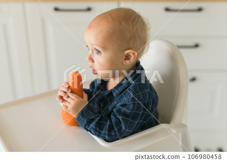 Happy baby sitting in high chair eating carrot in kitchen copy space. Healthy nutrition for kids. Bio carrot as first solid food for infant. Children eat vegetables. Little boy biting raw vegetable. 106980458