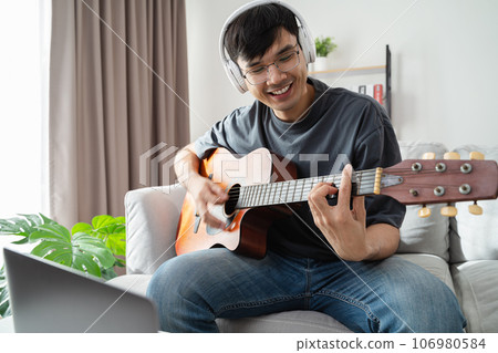 Young handsome man playing acoustic guitar sitting on the sofa in the living room at home. Young handsome man playing acoustic guitar sitting on the sofa in the living room at home. 106980584