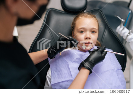 Closeup portrait of frightened cute little girl sitting in dental chair and looking at camera. Back view of unrecognizable female dentist holding in hands dental instruments during dental treatment. Closeup portrait of frightened cute little girl sitting in dental chair and looking at camera. Back view of unrecognizable female dentist holding in hands dental instruments during dental treatment. 106980917