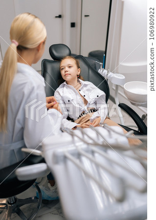 Vertical shot of sad little girl sitting on dental chair and complaining about pain visiting pediatric dentist. Prevention and prophylaxis of dental diseases. Concept of children teeth treatment. Vertical shot of sad little girl sitting on dental chair and complaining about pain visiting pediatric dentist. Prevention and prophylaxis of dental diseases. Concept of children teeth treatment. 106980922