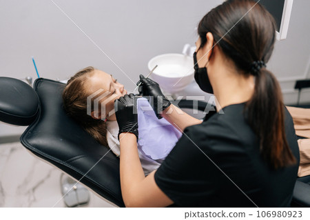 Side view of unrecognizable female dentist in gloves examining teeth of adorable little girl with dentistry equipment instrument lying on chair in dentistry clinic. Concept of children teeth treatment Side view of unrecognizable female dentist in gloves examining teeth of adorable little girl with dentistry equipment instrument lying on chair in dentistry clinic. Concept of children teeth treatment 106980923