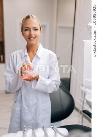 Vertical portrait of attractive female orthodontist doctor in white coat holding human jaws layout model with metal dental braces in modern dental clinic. Concept of stomatology, orthodontics. 106980926