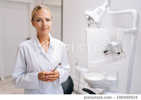 Portrait of attractive female dental doctor holding dental equipment for work and looking at camera in dentistry clinic with modern white interior. Concept of stomatology, dentistry and orthodontics. 106980928