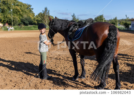 Horse riding school. Little children girls at group training equestrian lessons at outdoors ranch horse riding hall. Cute little beginner blond girl kid near beautiful brown horse 106982043