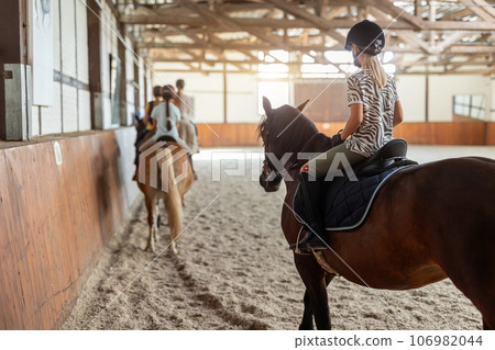 Horse riding school. Little children girls at group training equestrian lessons in indoor ranch horse riding hall. Cute little beginner blond girl kid in helmet sitting on brown horse horseback Horse riding school. Little children girls at group training equestrian lessons in indoor ranch horse riding hall. Cute little beginner blond girl kid in helmet sitting on brown horse horseback 106982044