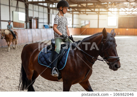 Horse riding school. Little children girls at group training equestrian lessons in indoor ranch horse riding hall. Cute little beginner blond girl kid in helmet sitting on brown horse horseback Horse riding school. Little children girls at group training equestrian lessons in indoor ranch horse riding hall. Cute little beginner blond girl kid in helmet sitting on brown horse horseback 106982046