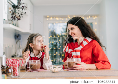 Happy smiling little daughter girl in Santa hat and her mom sitting on the decorated modern kitchen and drinking cocoa with marshmallows. Family holidays. Festive christmas mood. Selective focus Happy smiling little daughter girl in Santa hat and her mom sitting on the decorated modern kitchen and drinking cocoa with marshmallows. Family holidays. Festive christmas mood. Selective focus 106982843