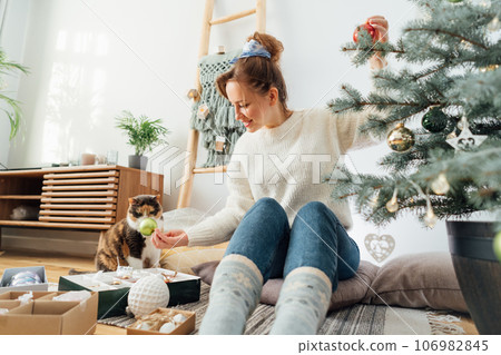 Young woman in cozy sweater decorating potted Christmas tree while her cat is watching her in light modern Scandinavian interior. Eco-friendly winter holidays. Domestic pet at home. Selective focus Young woman in cozy sweater decorating potted Christmas tree while her cat is watching her in light modern Scandinavian interior. Eco-friendly winter holidays. Domestic pet at home. Selective focus 106982845