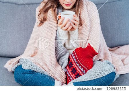 Woman freezes in wintertime. Young girl wearing warm woolen socks and wrapped into two blankets, holding a cup of hot drink and heating pad while sitting on sofa at home. Keep warm. Selective focus 106982855