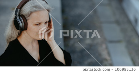 Head shot of a beautiful woman with gray hair sitting on concrete stairs in city, listening to music through wireless headphones. 106983136