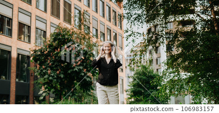 Portrait of a beautiful woman with gray hair listening to music through wireless headphones in city park. 106983157