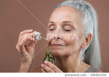 Close up of smiling beautiful senior woman holding skincare serum drops. Concept of beauty and skincare. 106983208