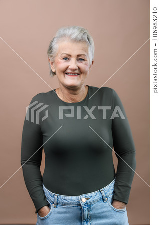 Portrait of smiling senior woman with hands in pockets in studio. 106983210