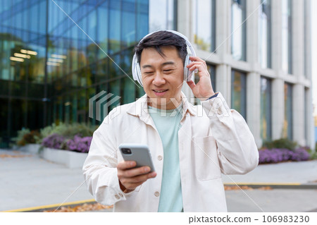 Asian young male student standing outside the campus wearing headphones and listening to music on the phone. He types with a smile and looks at the screen. Asian young male student standing outside the campus wearing headphones and listening to music on the phone. He types with a smile and looks at the screen. 106983230