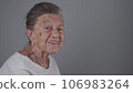Senior woman 90 years old smiles, laughs and shows a denture on a gray background. Elderly female with deep wrinkles and gray hair happy in a good mood studio portrait looking at camera 106983264