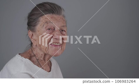 Senior woman 90 years old smiles, laughs and shows a denture on a gray background. Elderly female with deep wrinkles and gray hair happy in a good mood studio portrait looking at camera 106983264