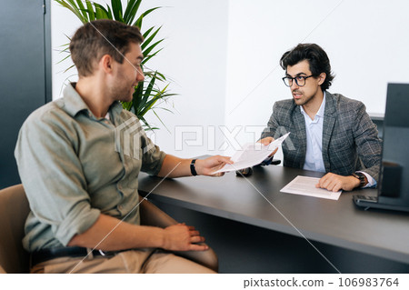 Smiling male job applicant arrives at interview, giving resume to serious hr recruiter wearing business suit. Confident manager receives application from client. Boss takes document for signing Smiling male job applicant arrives at interview, giving resume to serious hr recruiter wearing business suit. Confident manager receives application from client. Boss takes document for signing 106983764