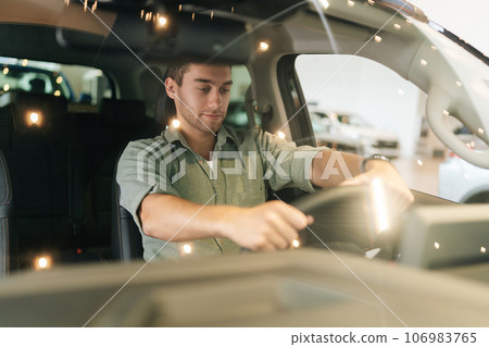 Portrait through windshield of pensive customer male client putting hand on steering wheel, looking down while trying new automobile at showroom. Satisfied young man client doing drive test of new car 106983765