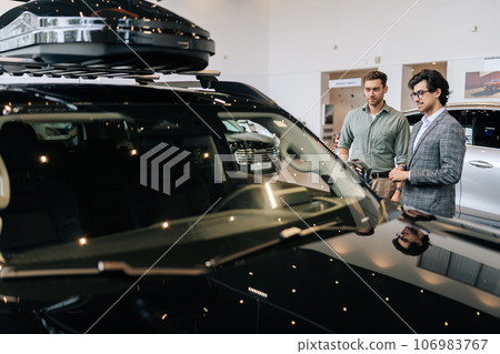 View from hood of vehicle to professional car dealer in business suit telling pensive male buyer about luxurious car in dealership. Pensive man client looking at auto. Concept of choosing new car. View from hood of vehicle to professional car dealer in business suit telling pensive male buyer about luxurious car in dealership. Pensive man client looking at auto. Concept of choosing new car. 106983767