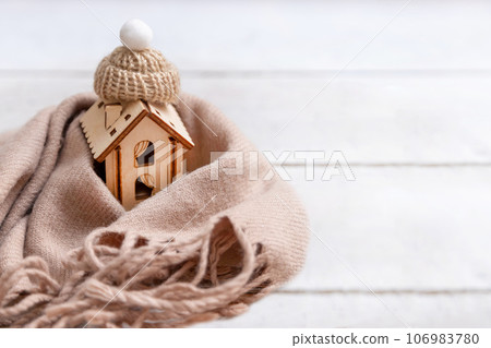 A small house, wrapped in a scarf with a hat, stands on a wooden background. A small house, wrapped in a scarf with a hat, stands on a wooden background. 106983780