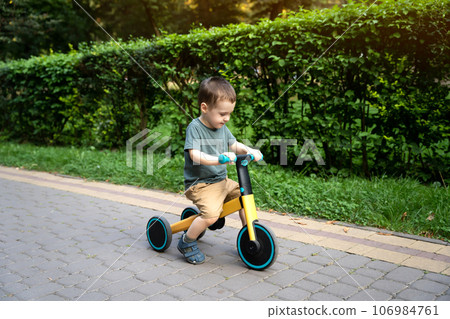 A smiling toddler boy of two or three years old rides a bicycle or balance bike in a city park on a sunny summer day. Toddlerhood and childhood concept. Selective focus 106984761