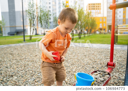 Little toddler boy playing with toys on pebbles on the playground. Boy two or three years old. Toddlerhood childhood concept. Residential complex of apartment buildings. 106984773