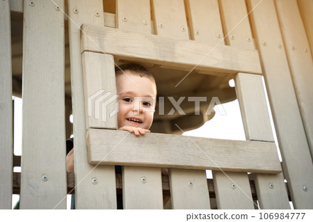 A happy smiling toddler boy of two or three years looks out of a window on a wooden playground. Toddlerhood and childhood concept. Active lifestyle 106984777