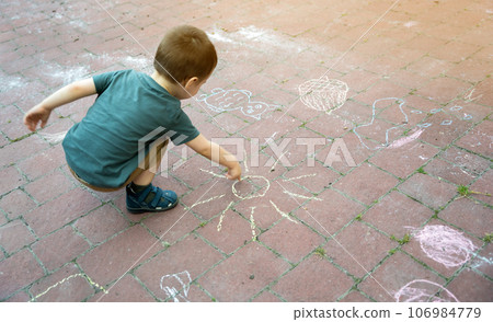 Little toddler boy two or three years old draws sun with crayons in the park on tiles. Playtime for children. Children's games. Selective focus 106984779
