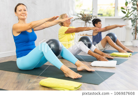 Middle-aged woman practicing fitness with softball between legs in light training room Middle-aged woman practicing fitness with softball between legs in light training room 106987608
