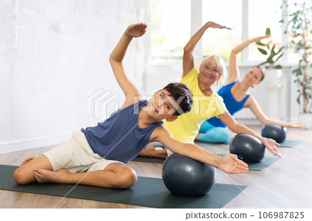 Preteen boy practicing side bend lotus pose of yoga with softball in light fitness room Preteen boy practicing side bend lotus pose of yoga with softball in light fitness room 106987825