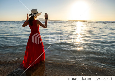 Young woman wearing long red dress and straw hat standing in sea water at the beach enjoying view of rising sun in early summer morning. 106990801