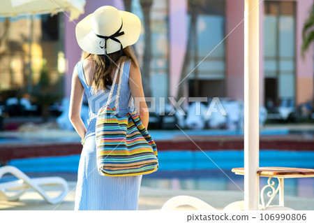 Young woman wearing light summer dress and yellow straw hat holding fashionable shoulder bag standing outside hotel on summer sunny day. Young woman wearing light summer dress and yellow straw hat holding fashionable shoulder bag standing outside hotel on summer sunny day. 106990806