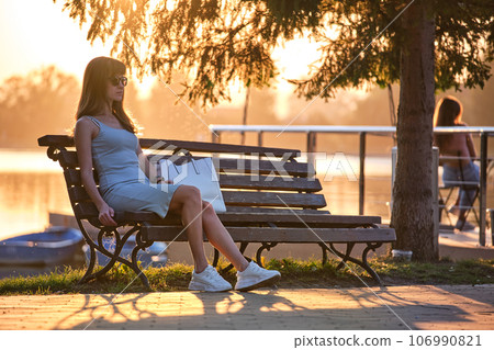 Young woman in summer dress sitting relaxed on lake side bench on warm evening. Wellness resting from everyday rush concept. 106990821