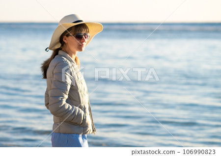 Young woman in straw hat and a dress standing alone on empty sand beach at sea shore. Lonely tourist girl looking at horizon over calm ocean surface on vacation trip. 106990823