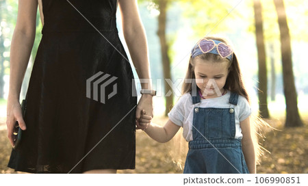Young mother and her small daughter with long hair walking together holding hands in summer park. 106990851