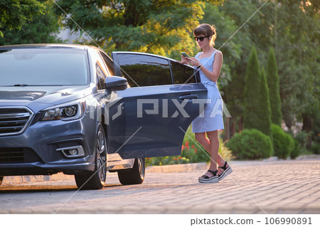 Young female driver standing near her vehicle browsing cellphone on a city street in summer. Young female driver standing near her vehicle browsing cellphone on a city street in summer. 106990891